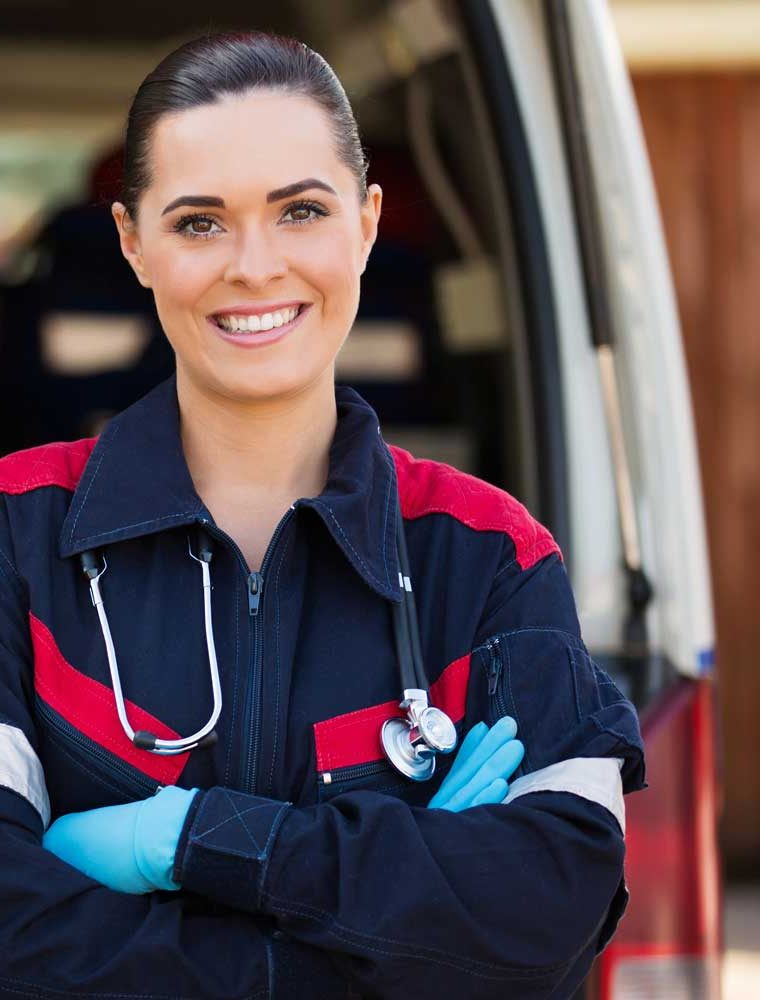 A smiling paramedic in uniform with a stethoscope, standing at the open back of an ambulance